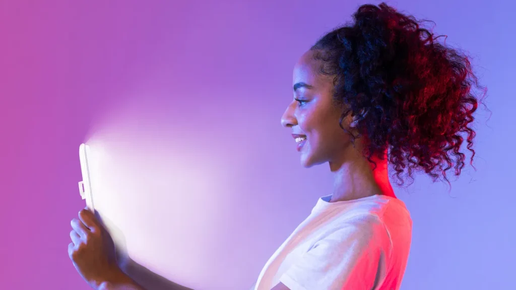 a woman with brown curly hair looking at a bright screen light with a purple and pink background